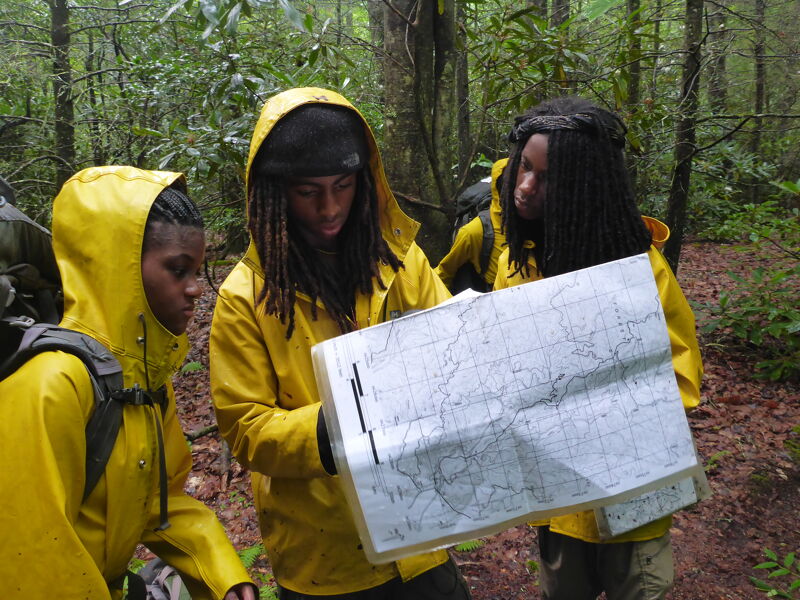 Three young hikers wearing yellow raincoats huddle together in a lush, green forest, carefully studying a large map. The hikers appear to be focused on navigation, possibly determining their route or current location. The dense foliage and overcast lighting suggest they are deep in the wilderness, likely on a challenging trek. Their expressions convey a sense of determination and teamwork as they navigate the trail.